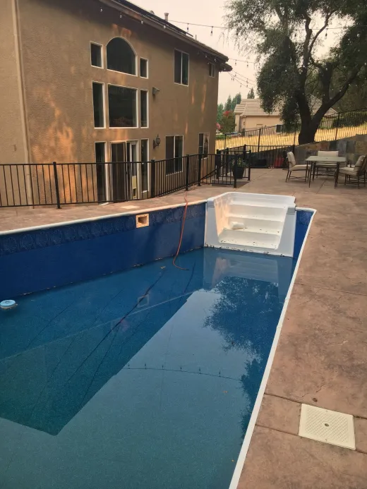 Backyard pool with blue tile interior next to a two-story beige house and patio furniture.