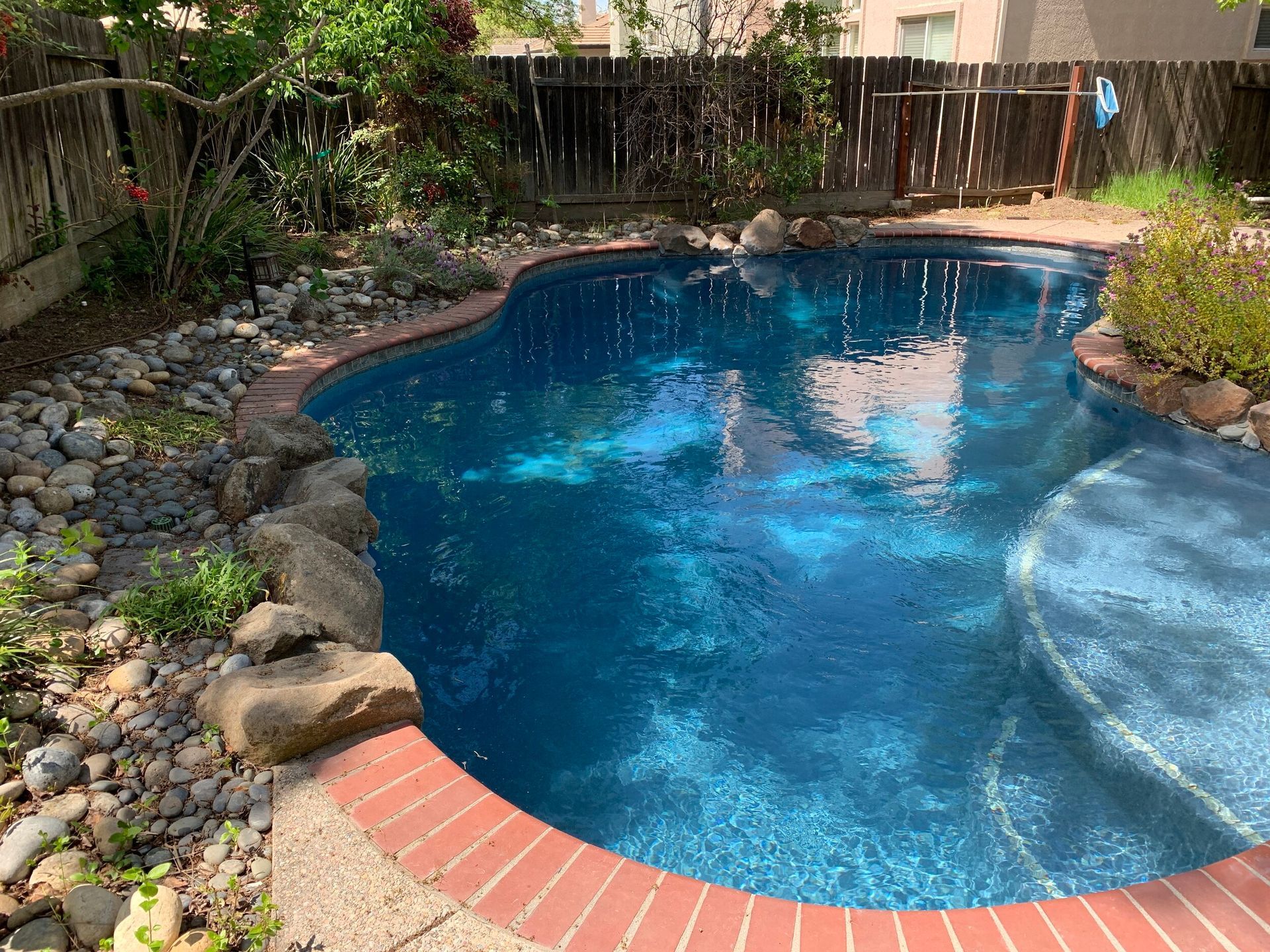 A swimming pool with blue water surrounded by brick and rocks, backyard setting.
