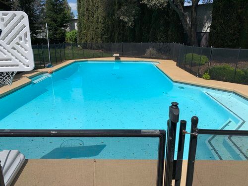 A turquoise swimming pool with a basketball hoop, surrounded by a black fence and concrete deck.