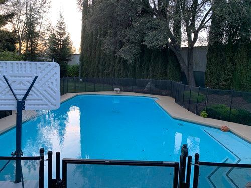 Swimming pool with basketball hoop and surrounding fence, trees in background, during daytime.