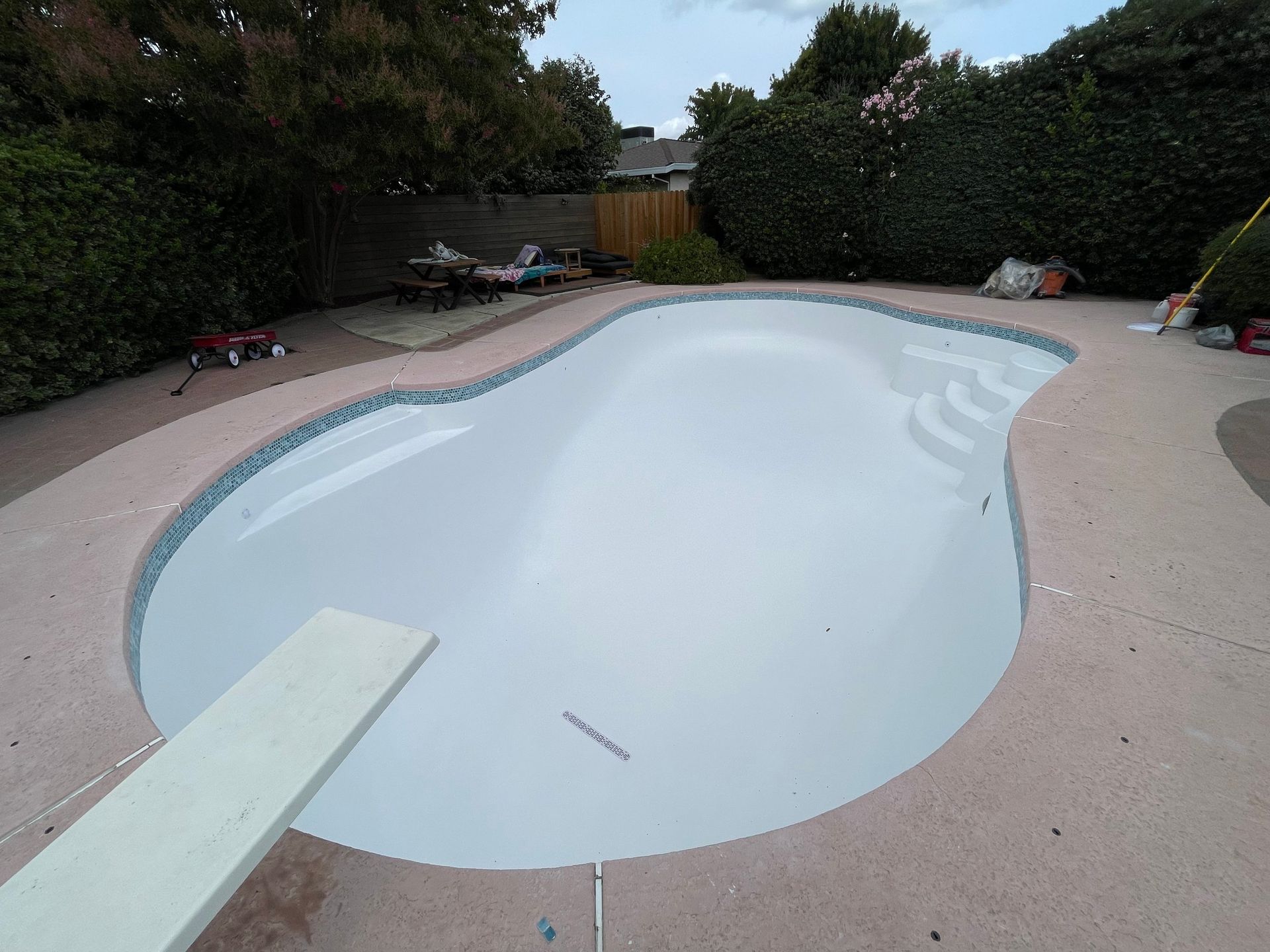 Empty kidney-shaped swimming pool with diving board, surrounded by concrete deck and landscaping.