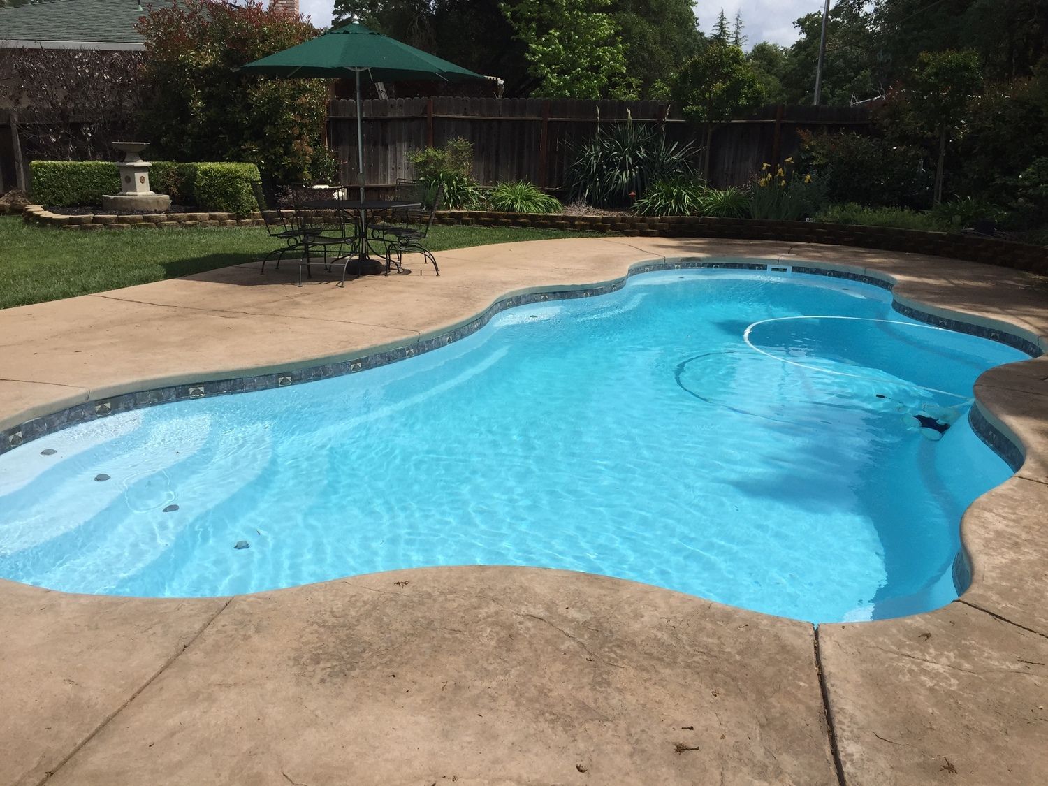 Swimming pool with blue water and surrounding concrete patio, table and umbrella on the grass.