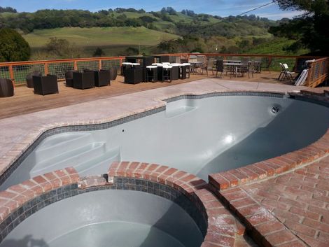 An empty pool and hot tub next to a brick patio with outdoor furniture, set against a green hillside.