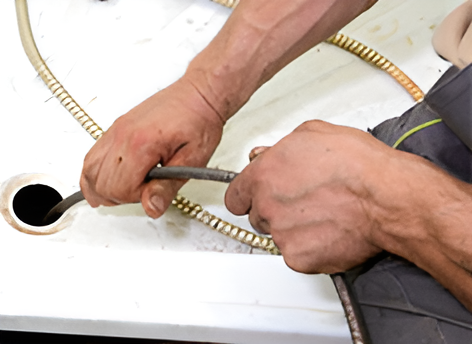 A person's hands are using a plumbing snake to unclog a drain in a white sink. They're gripping the coiled cable and inserting it into the drain opening.