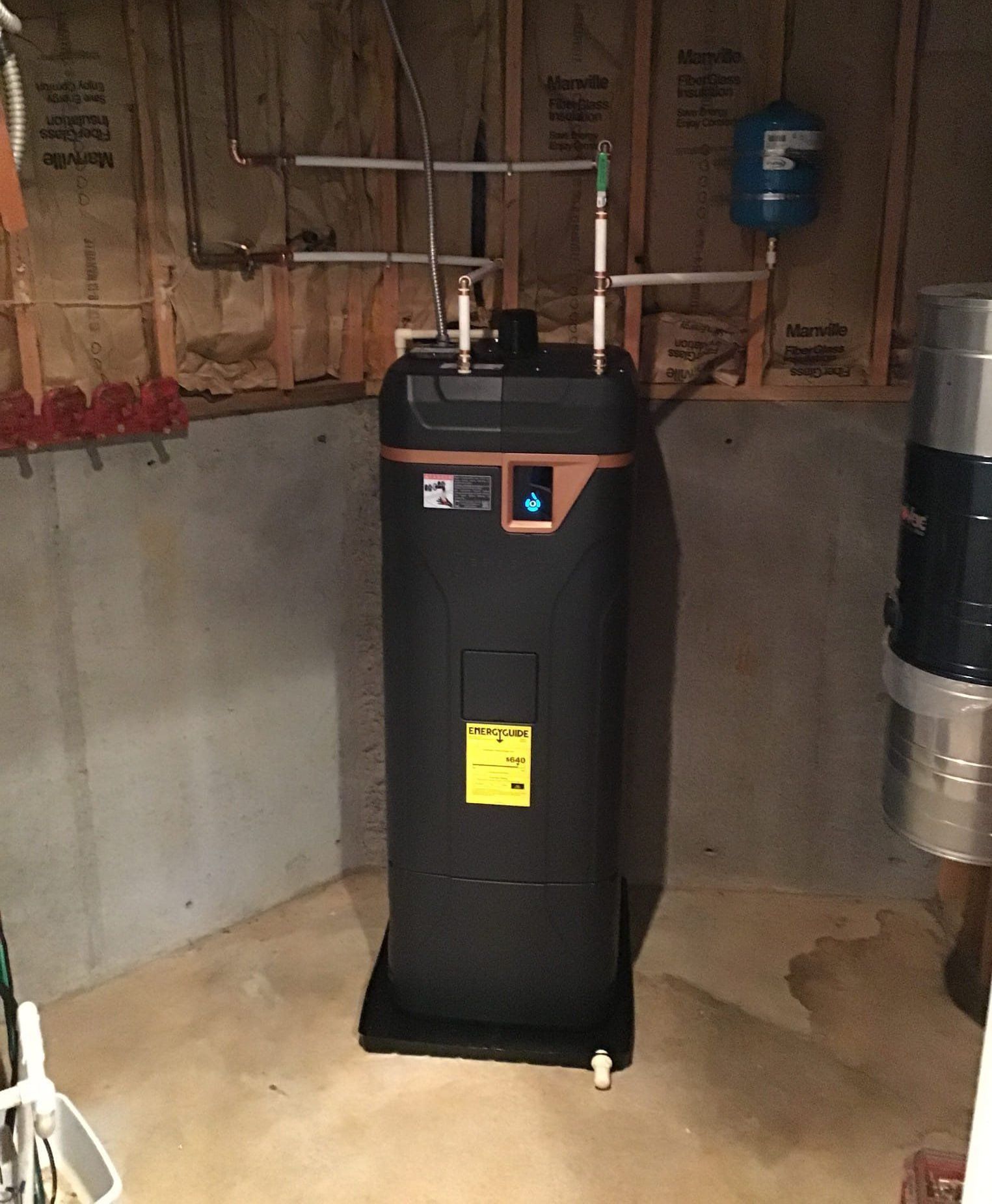 Black water heater in a basement corner, with pipes and a blue tank visible.