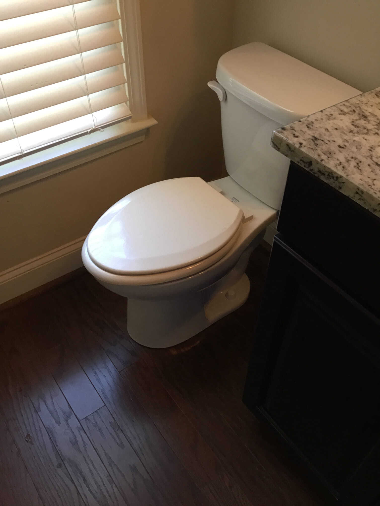 Toilet in a bathroom corner, next to a window with blinds and a dark cabinet.