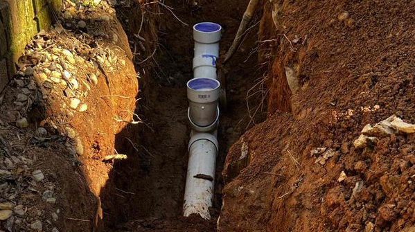 Trench in dirt with white PVC pipes and fittings.