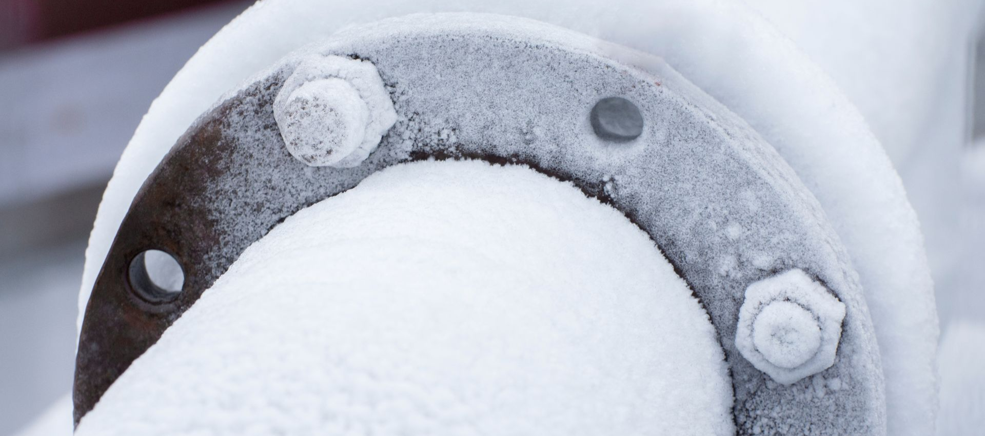 A close-up of a snow-covered industrial pipe flange with bolts and a hole, outdoors.