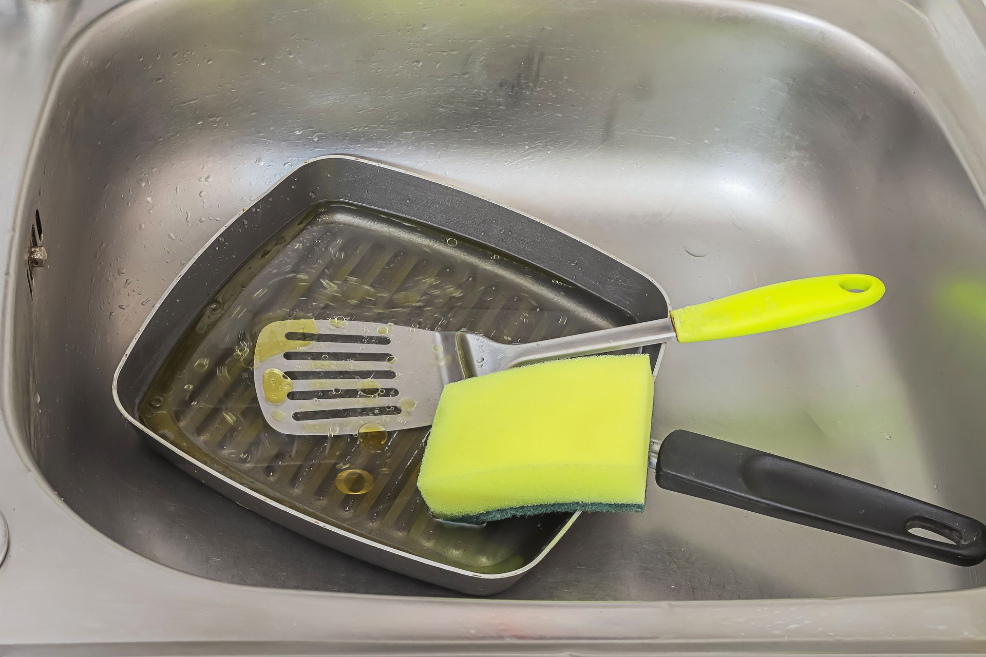 Grill pan, spatula, and sponge in a stainless steel kitchen sink.