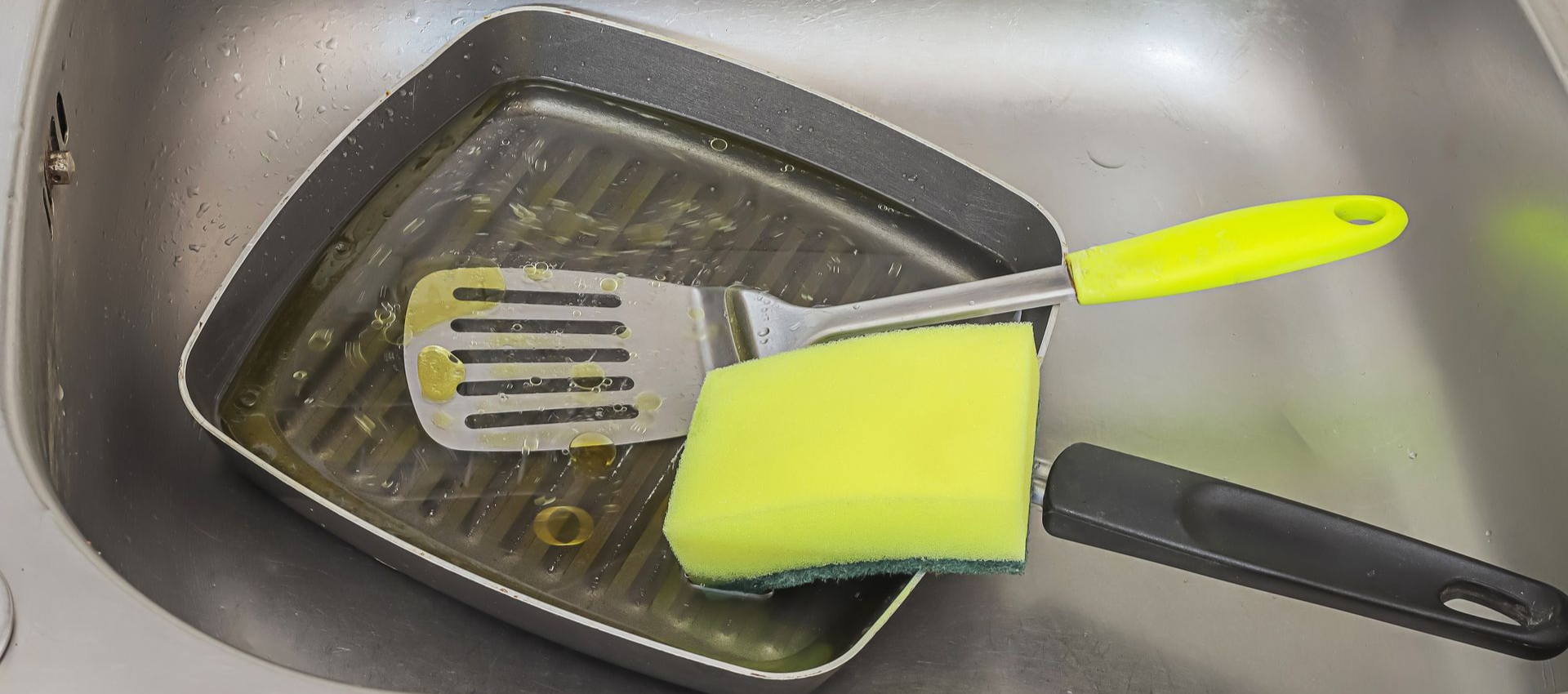 A dirty grill pan with a spatula and sponge in a sink.