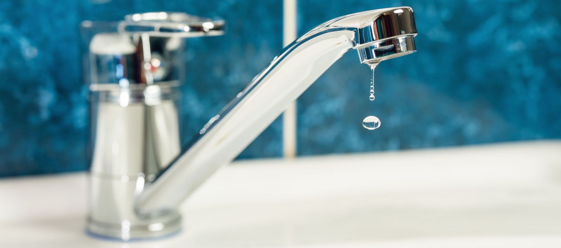 Chrome faucet with a water droplet falling, on a white surface, against blue tile.