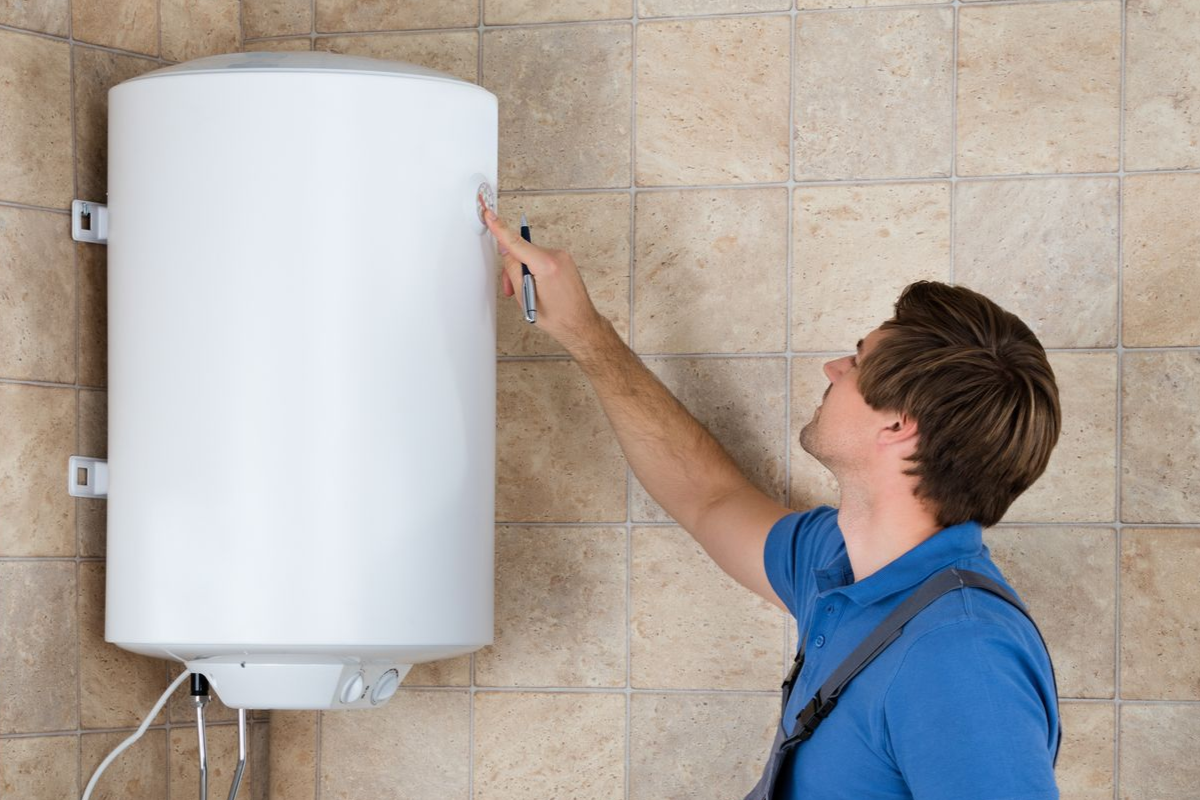 A male plumber inspecting a white water heater attached to a tile wall in a bathroom.