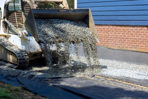 A skid steer loader dumps gray gravel onto a black landscaping fabric near a brick and blue-sided building.