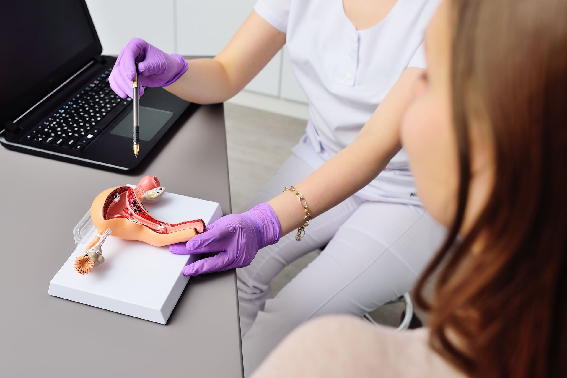 Doctor in purple gloves pointing to anatomy model during consultation with patient on laptop nearby