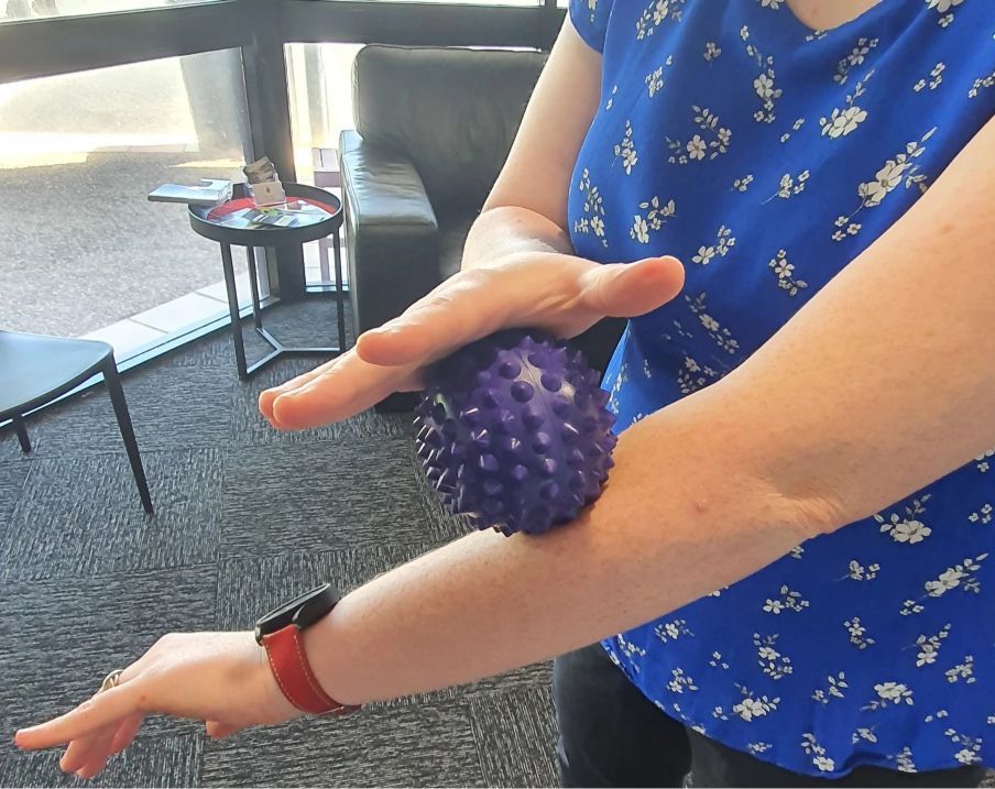 A Woman in a Blue Shirt is Holding a Purple Ball on Her Elbow — Anderson Family Chiropractic Health Centre in Mount Pleasant, QLD
