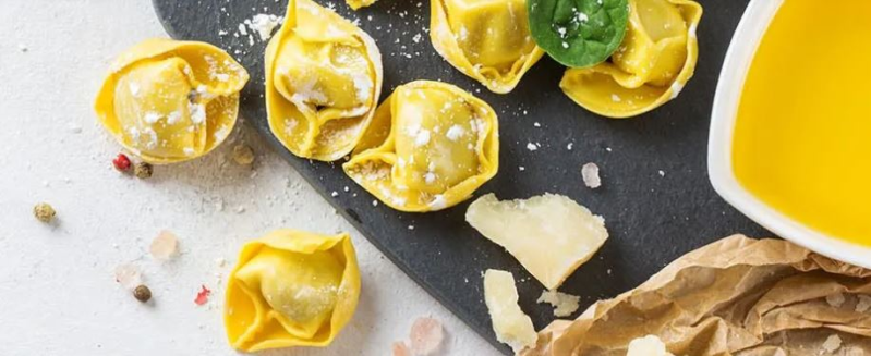 Casoncelli su una tavola di ardesia con una ciotola di salsa e formaggio grattugiato, su una superficie bianca.