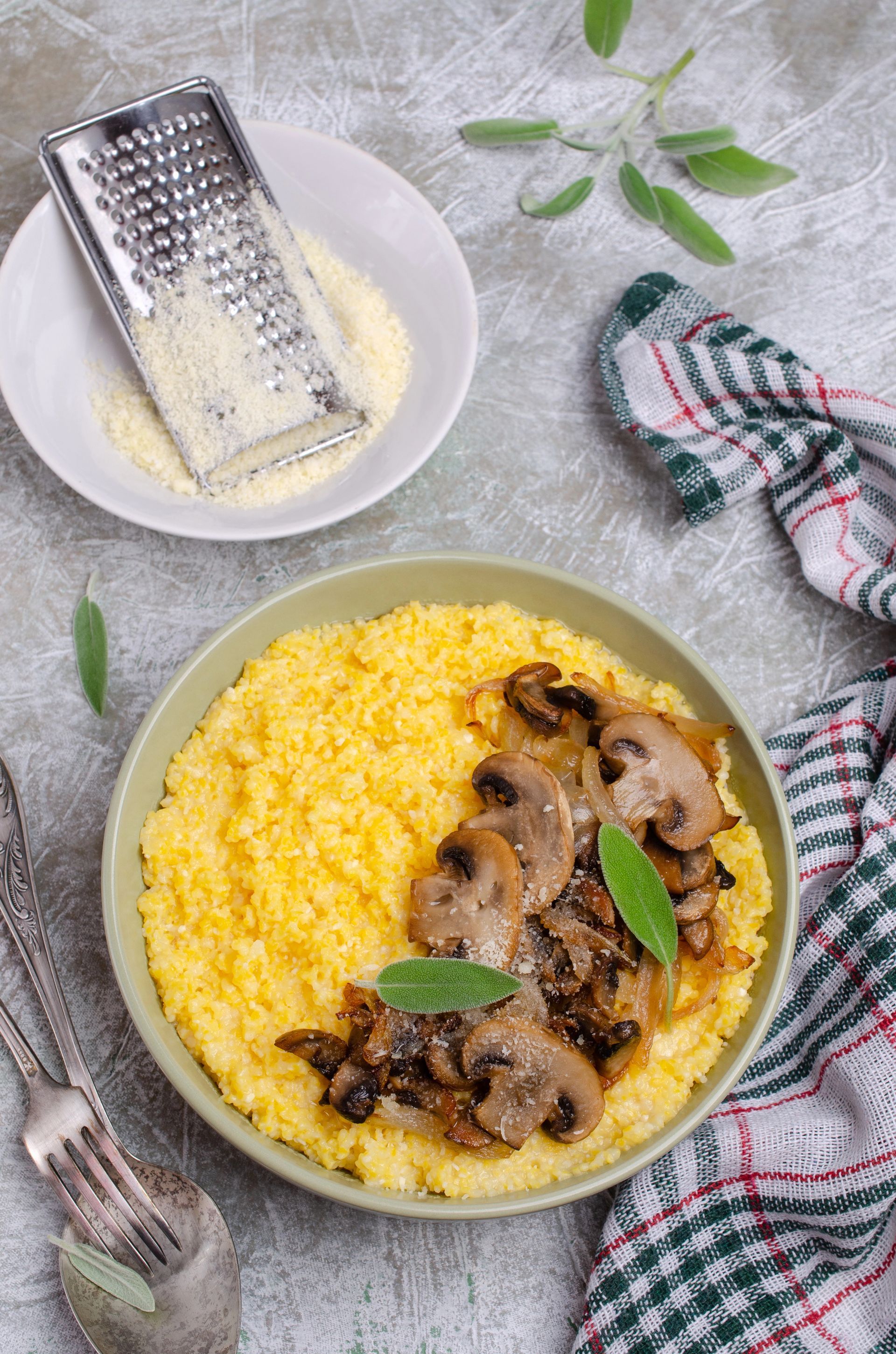 Polenta con funghi saltati in una ciotola, parmigiano grattugiato, salvia e un canovaccio a quadretti.