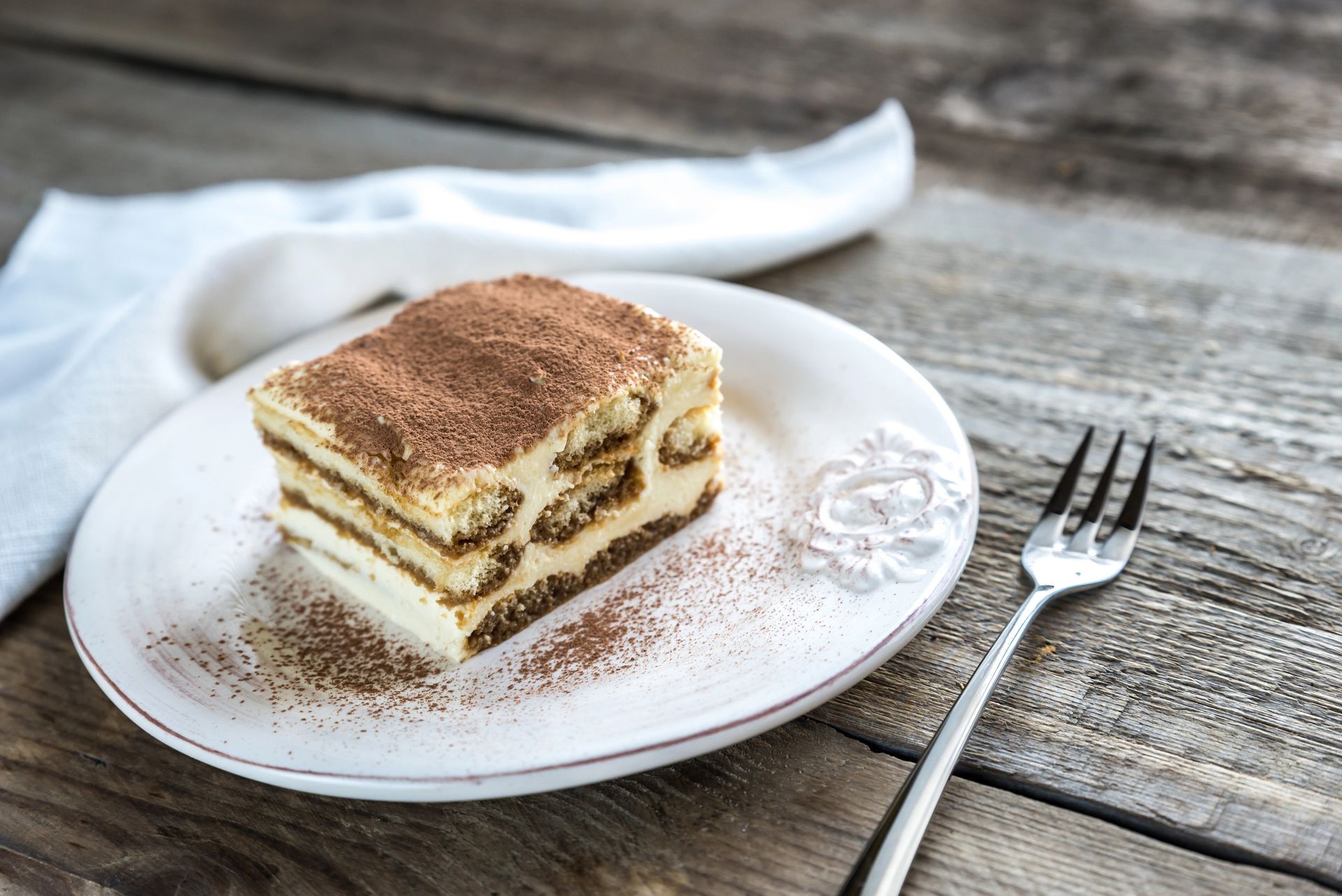 Tiramisù servito su un piatto bianco con una forchetta, su un tavolo di legno. Dessert spolverato con cacao in polvere.