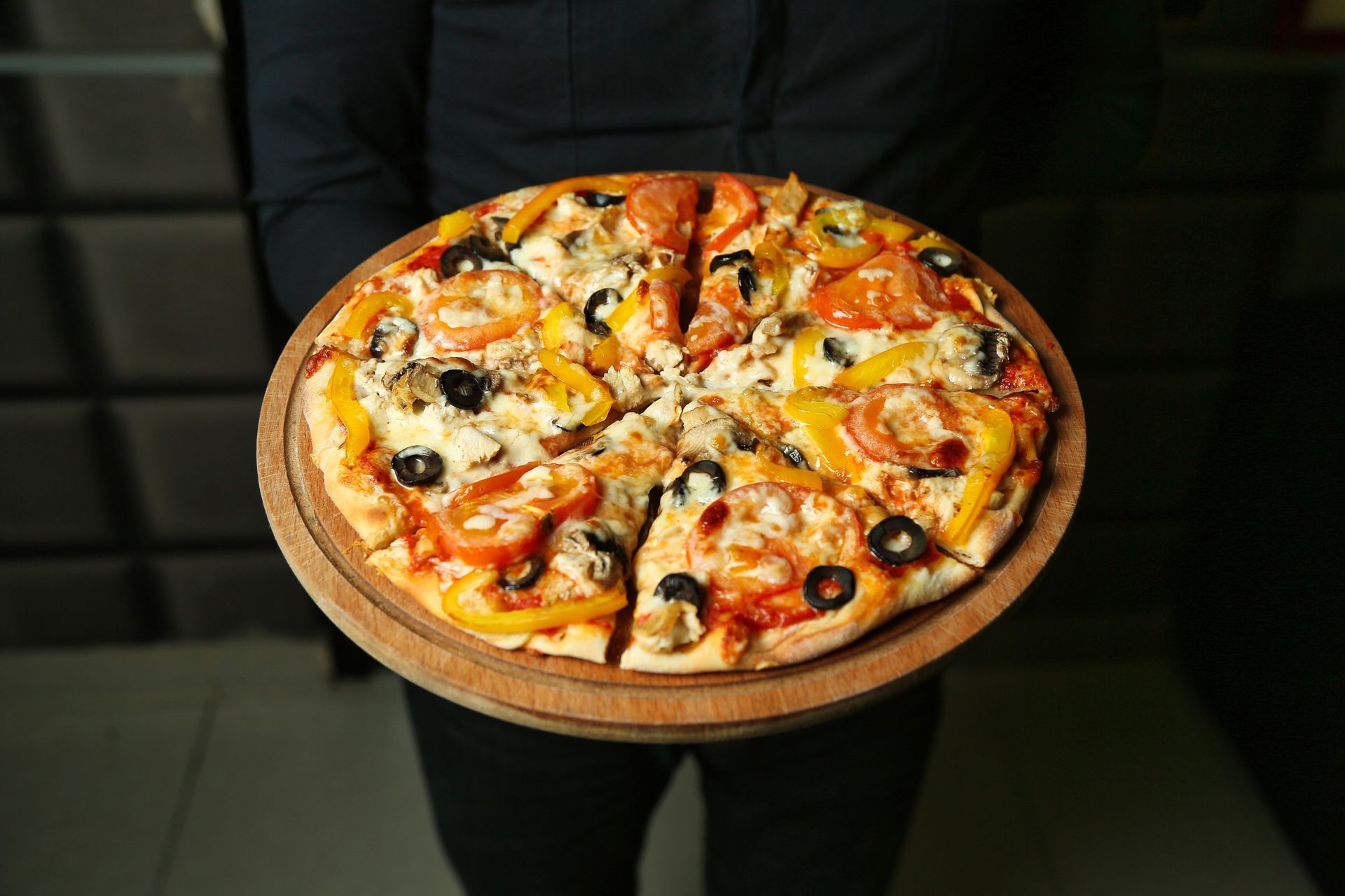 Pizza su un tagliere di legno tenuto da una persona. La pizza è condita con olive, peperoni e pomodori.