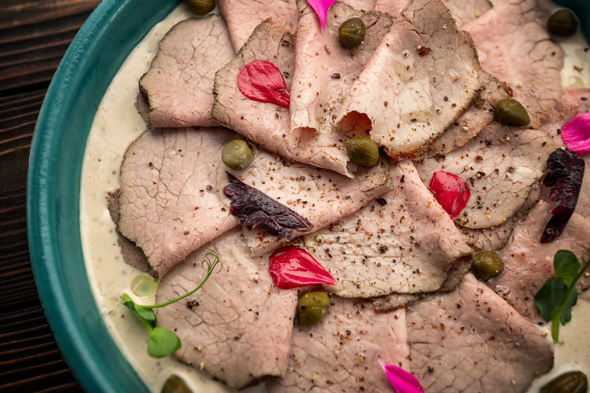 Vitello tonnato tagliato sottile con salsa cremosa, capperi e peperoni rossi su un piatto verde acqua.