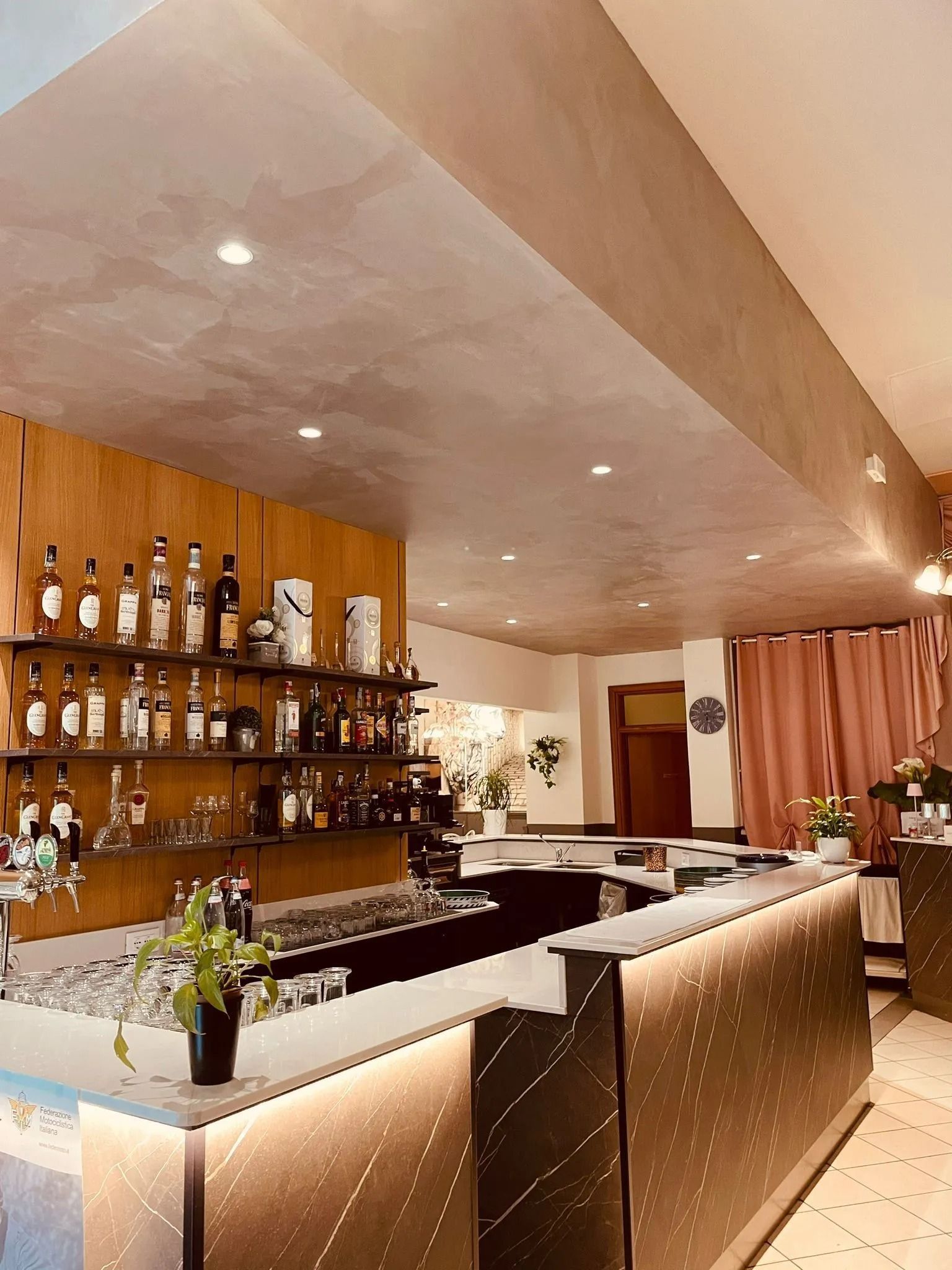 Interno del bar con bottiglie di liquore sugli scaffali, bancone illuminato, posti a sedere e tende rosa.