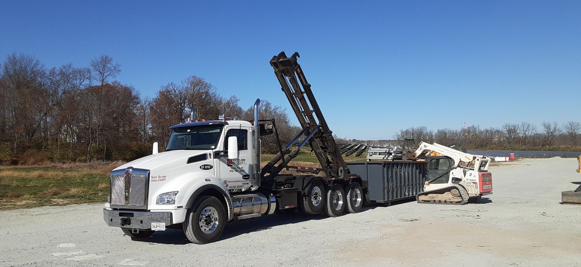 A white roll-off dump truck with a raised hydraulic arm loading a dumpster next to a small skid steer on a gravel lot.