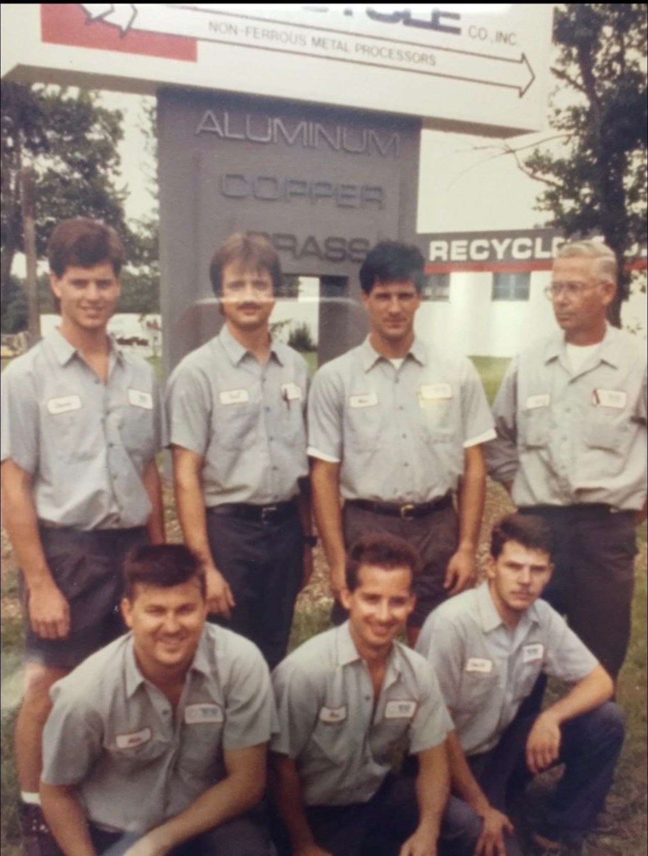 Seven employees in matching uniforms pose in front of a metal recycling company sign.