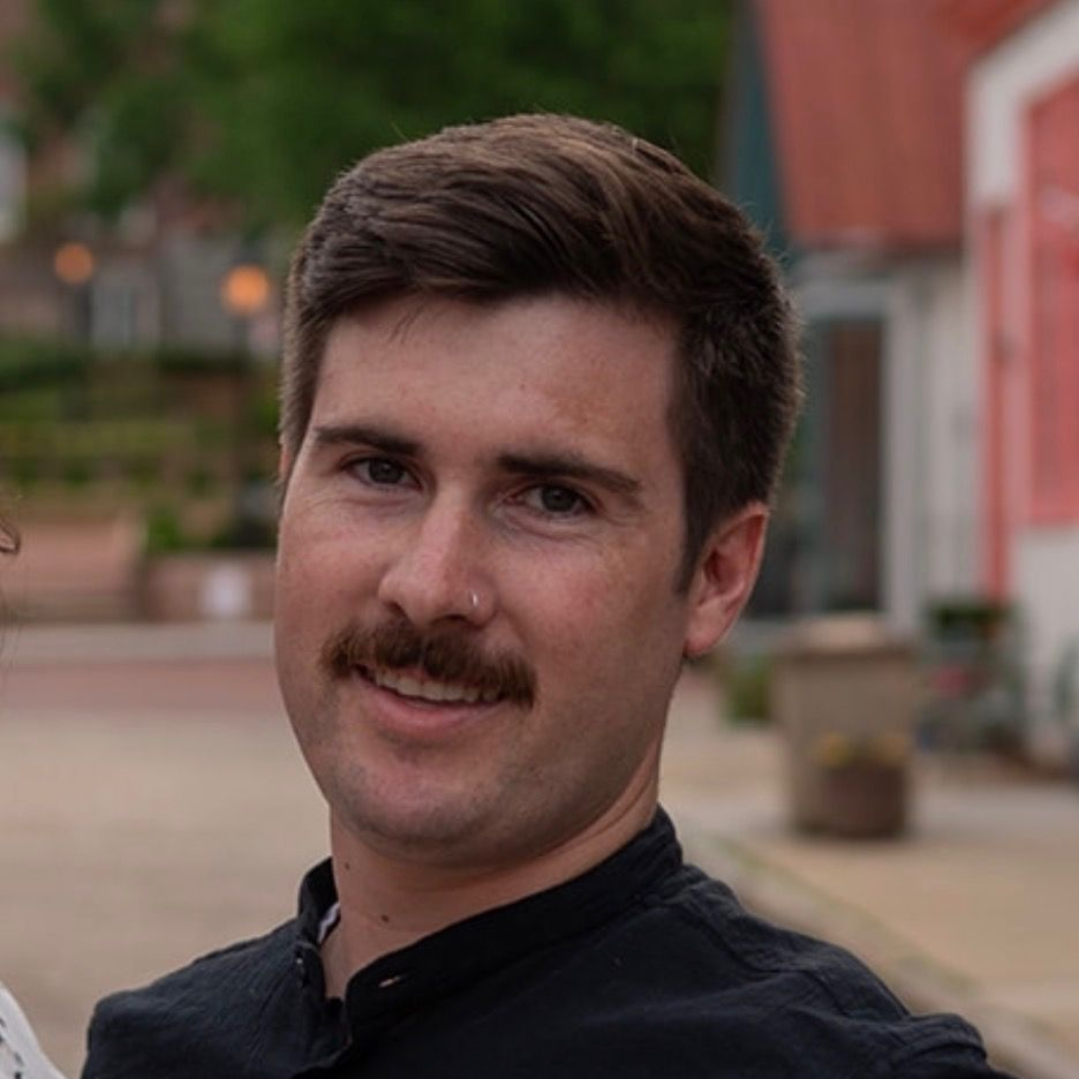 A smiling man and woman stand in front of large, stacked, metal-bound cubes of compressed aluminum cans outdoors.