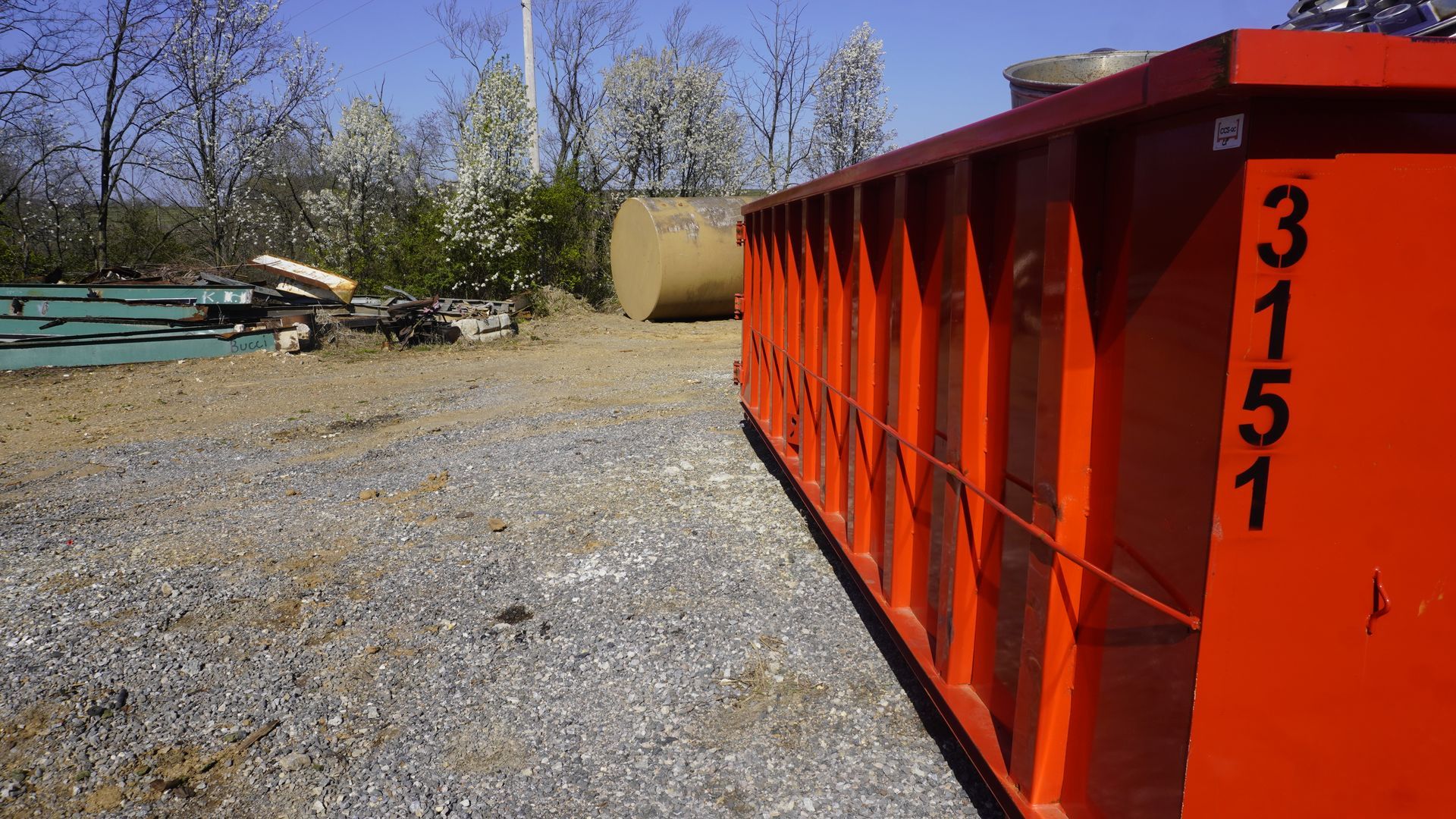Orange dumpster in gravel lot with trees and equipment in the background.