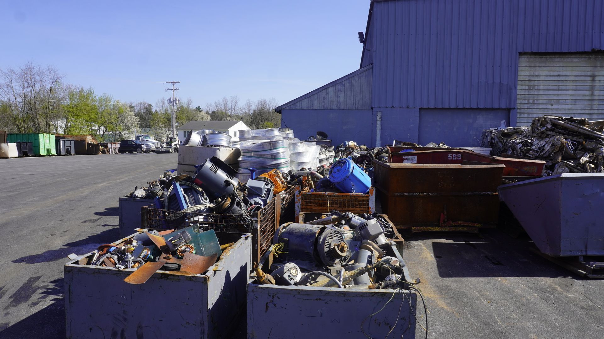 Outdoor junkyard with bins of scrap metal and debris beside a large blue industrial building