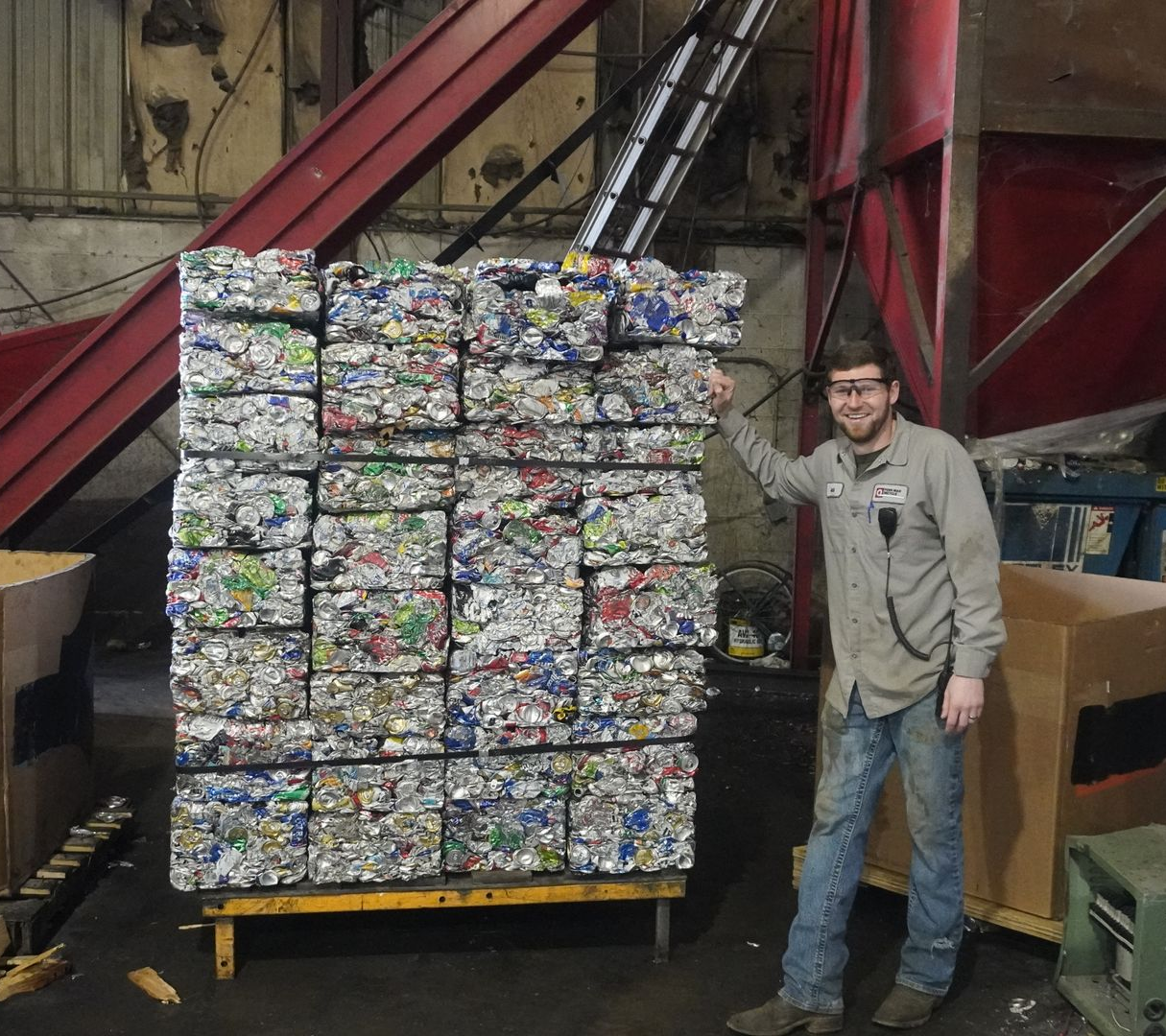 Man standing beside a pallet stacked with crushed cans in an industrial warehouse