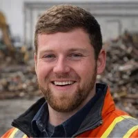 Three people, including two wearing gray work shirts and one in a pink shirt, standing outdoors in front of a vehicle.