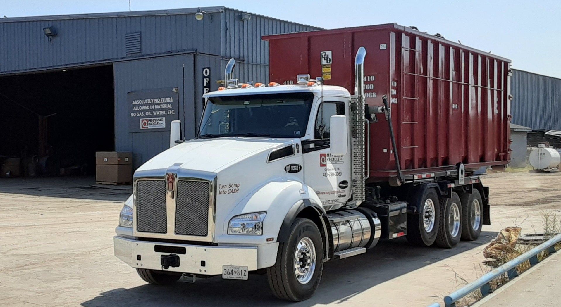 A white Kenworth roll-off dump truck with a large, empty red dumpster parked in an industrial lot.