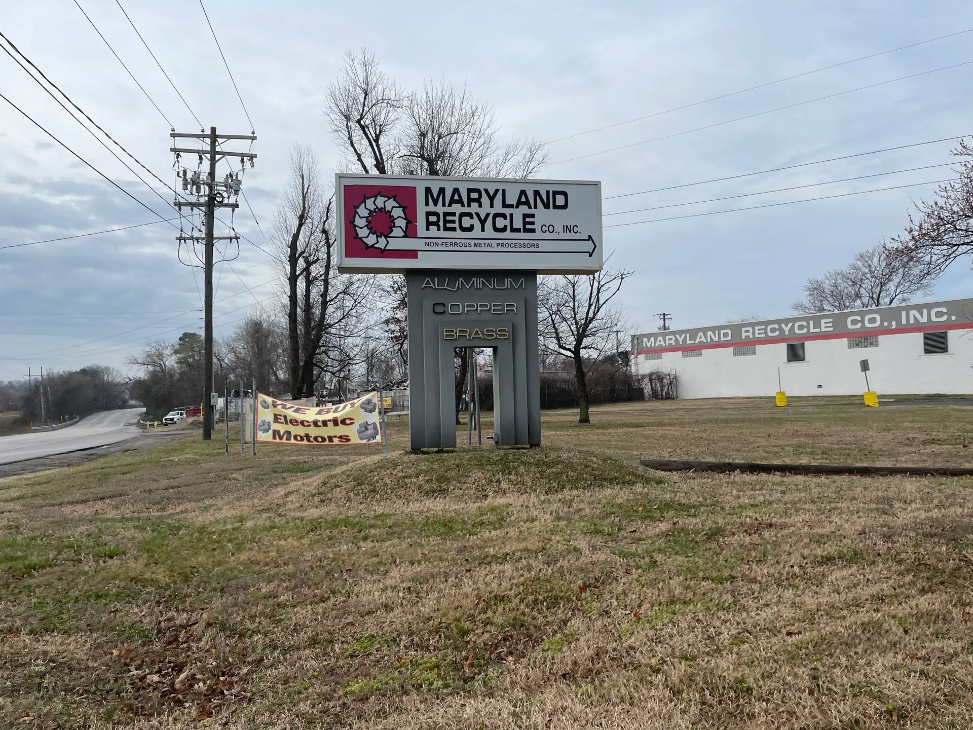 A sign for Maryland Recycle Co. stands on a grassy hill near a building marked Maryland Bicycle Co., Inc. under a cloudy sky.