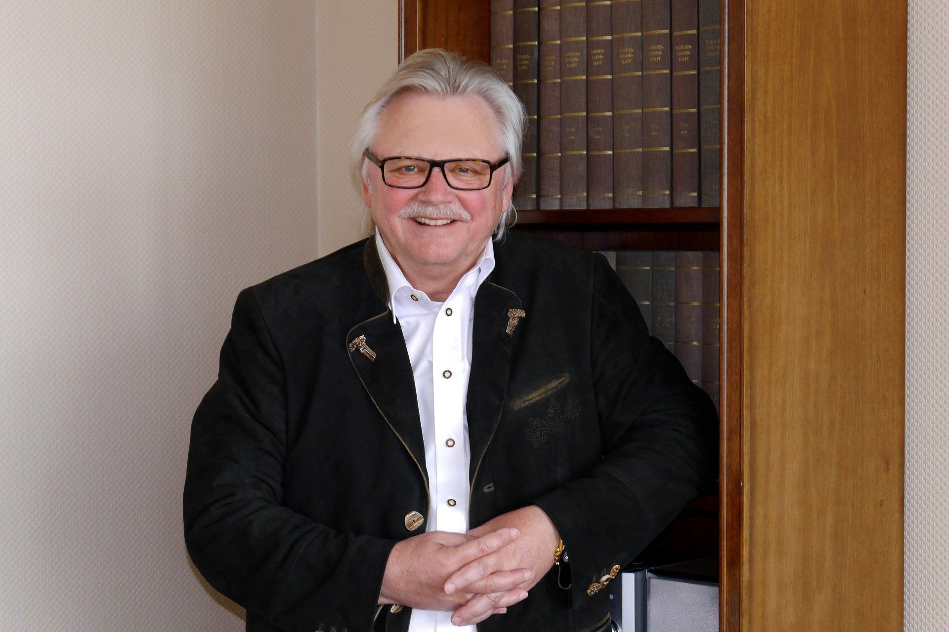 Ein Mann mit Brille und schwarzer Jacke steht vor einem Bücherregal.