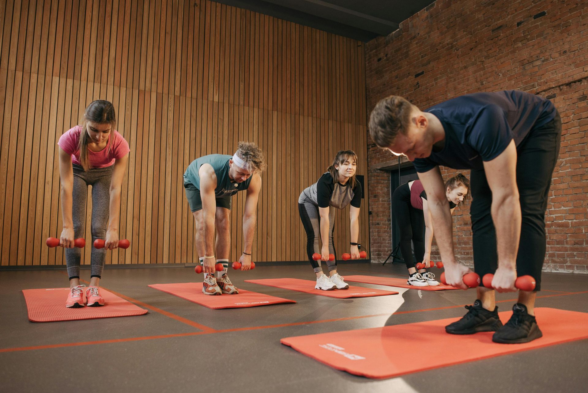 People exercising on mats, bending over with dumbbells in a studio with wooden walls and red accents.