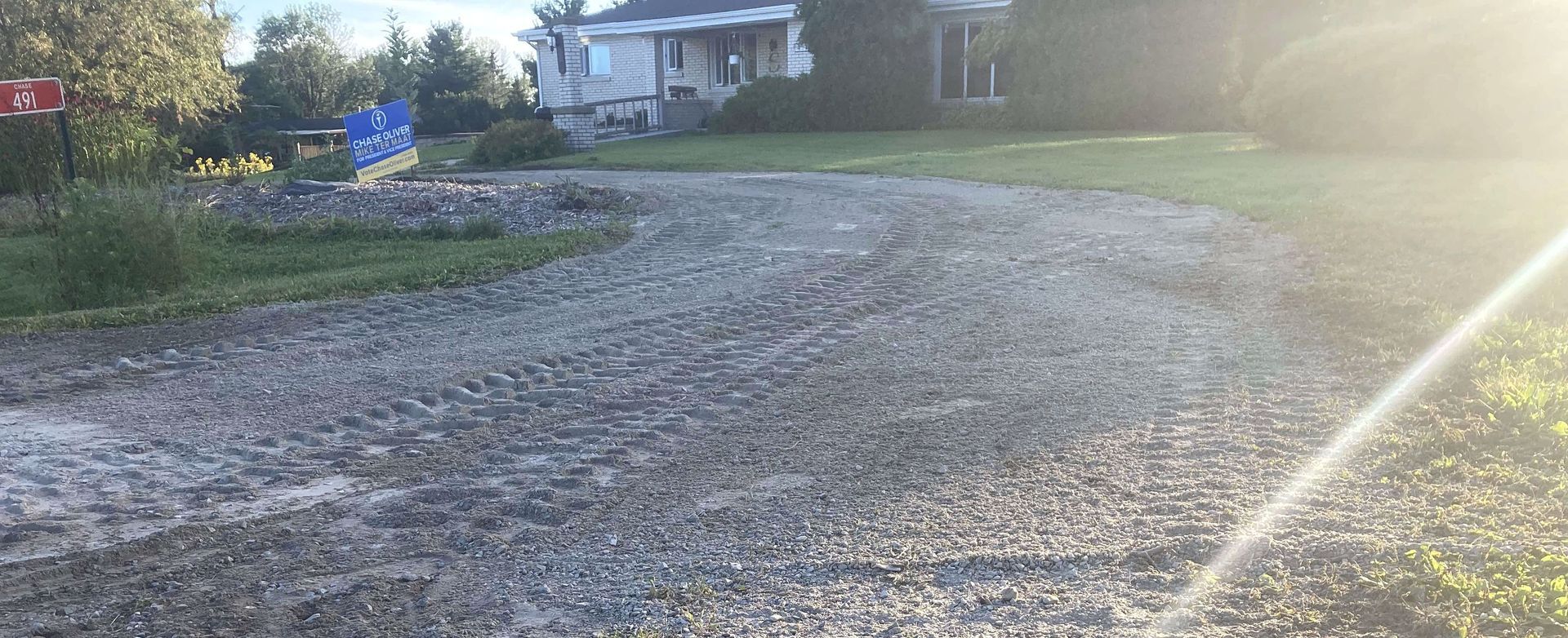 A gravel road leading to a house with the sun shining through the trees.