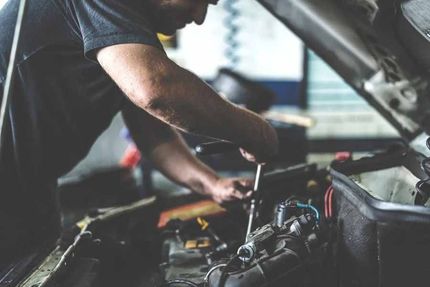 A mechanic leaning over an open car hood, using a tool to work on the engine in a repair shop.