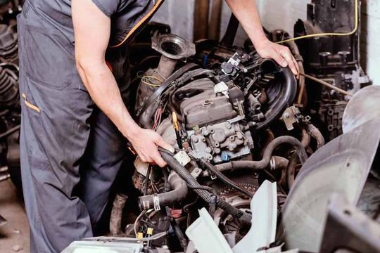 A person in a gray uniform works on a car engine in a cluttered auto shop.