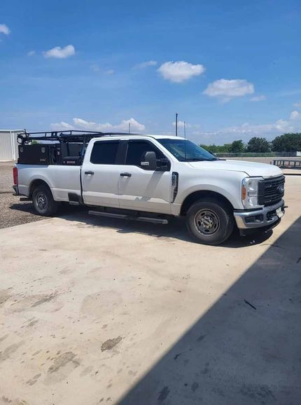 A white Ford Super Duty crew cab pickup truck with a black utility rack, parked on a concrete lot under a blue sky.