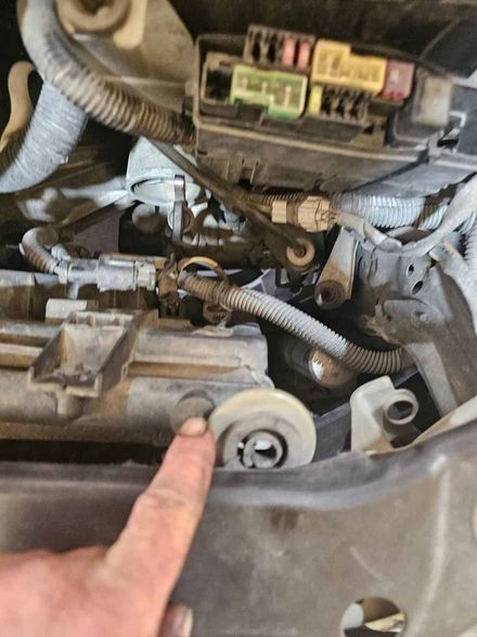 A person's finger points to a rubber mounting bushing on a car radiator near the engine fuse box.