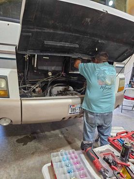 A person in a light blue t-shirt works on the engine of a large, beige-fronted vehicle with its hood propped open.