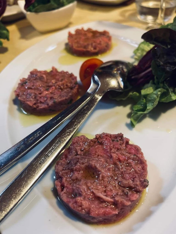 Tre porzioni di tartare di manzo su un piatto, guarnite con lattuga, un pomodoro e olio d'oliva. Le forchette sono incrociate sul piatto.