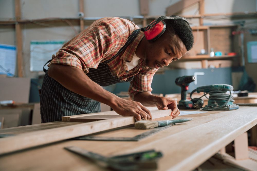 A young man wearing ear muffs is working on a piece of wood in a workshop.