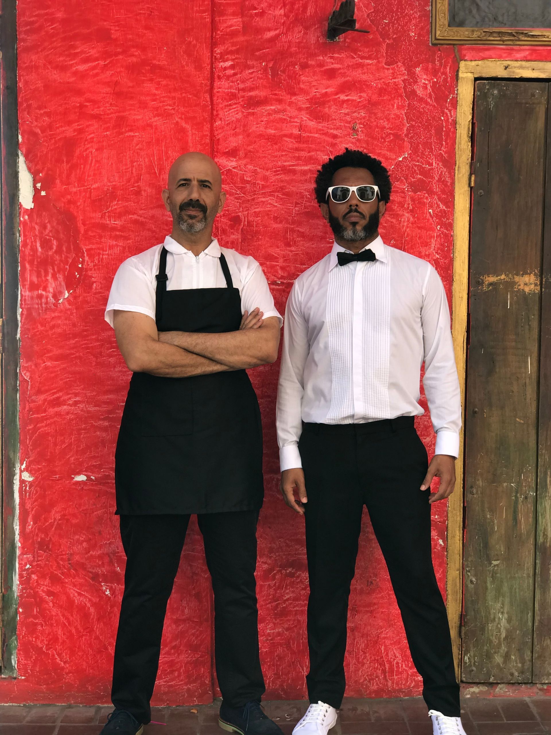Dos hombres de pie uno al lado del otro frente a una pared roja.