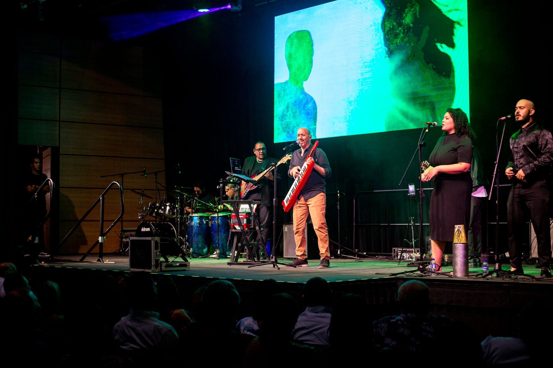 Un grupo de personas toca instrumentos en un escenario frente a una pantalla grande.