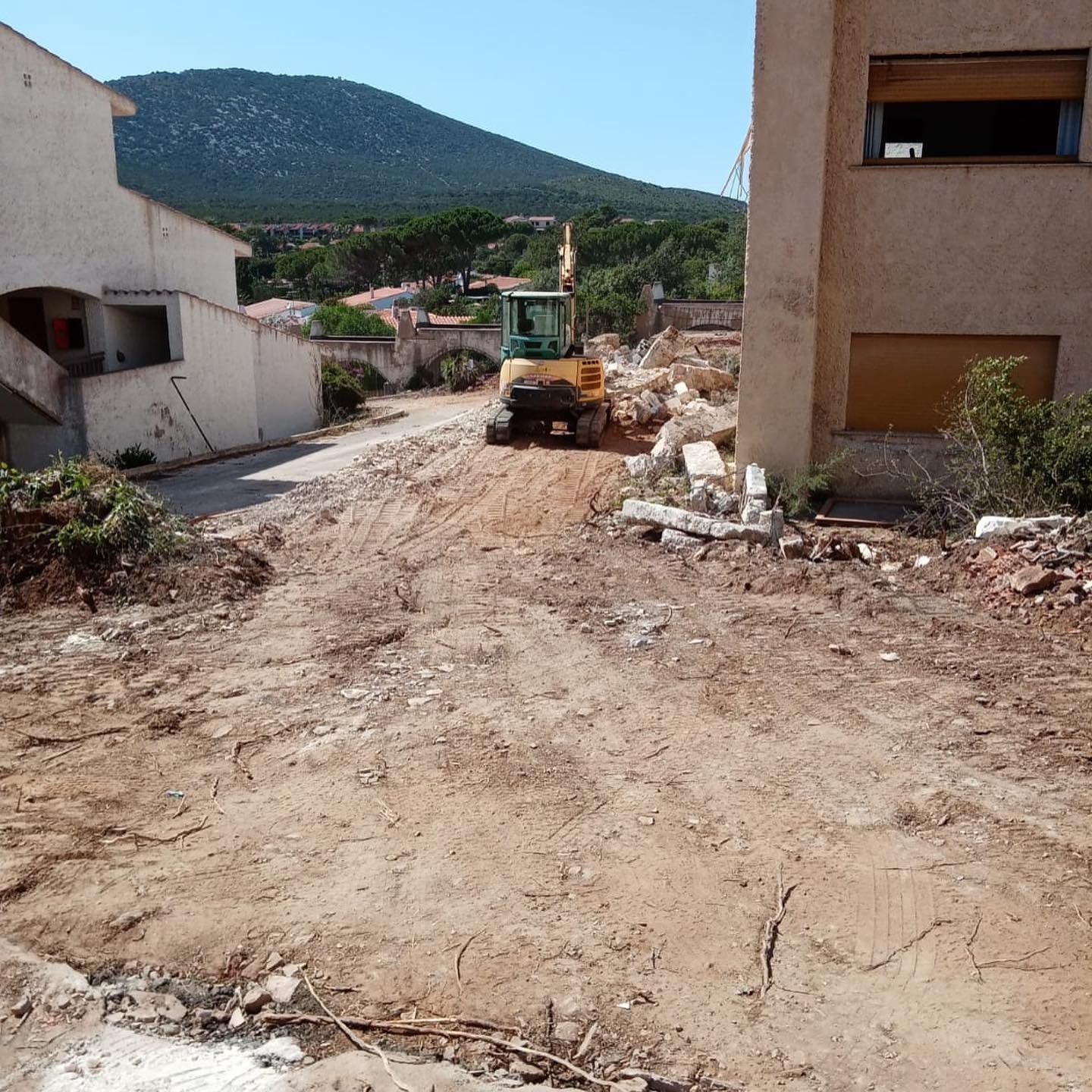 Un escavatore su una strada sterrata tra due edifici, montagne sullo sfondo. Cantiere edile.