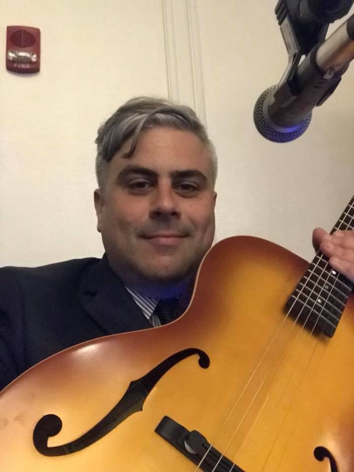 A man in a suit is holding a guitar in front of a microphone