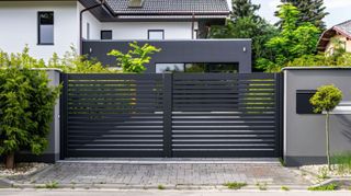Black, horizontal slat driveway gates with adjacent gray walls and house in background.