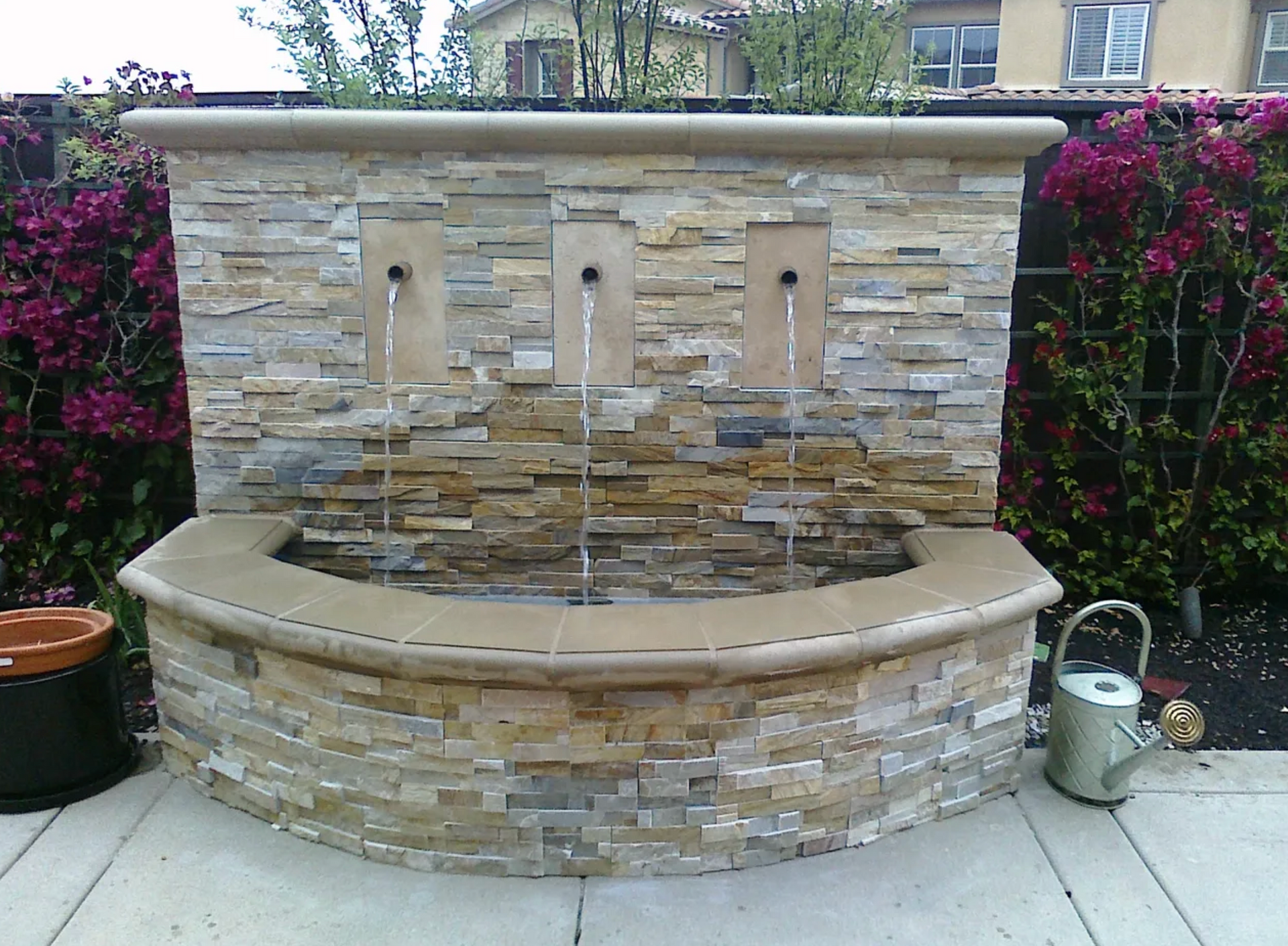 A water fountain is surrounded by flowers and a watering can